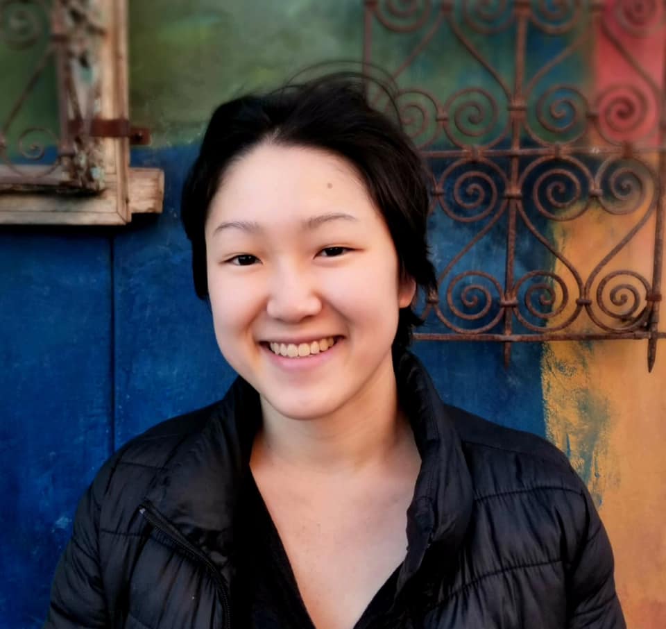 A headshot of Annie, a smiling white person with long straight brown hair, brown eyes, black glasses, and a maroon sweater