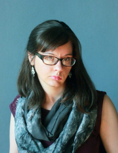 a white woman with dark brown hair, she wears glasses, jade-colored earrings, and has light pink lipstick. She wears a blue, faced infinity scarf that matches the blue background