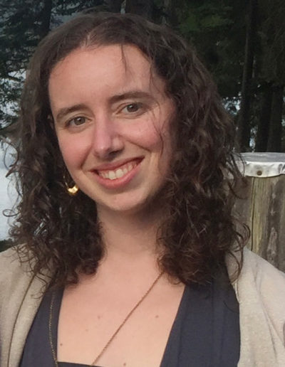 a white woman with curly brown hair smiling, the bottom half of a golden hoop earing is visible on her right ear. She wears plain colored clothes and is in a forest.