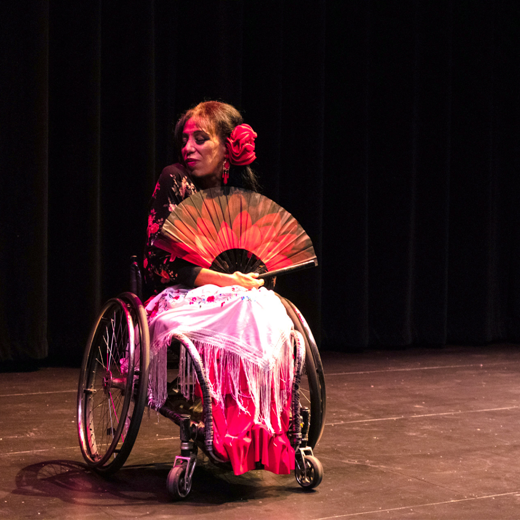 On a bare stage, a Latina woman wearing long, silky clothes on a wheelchair. She looks coyly to the side, holding a red and black fan over her chest with one hand. She wears a large red paper flower in her hair that matches her earrings, lipsticks, and outfit.