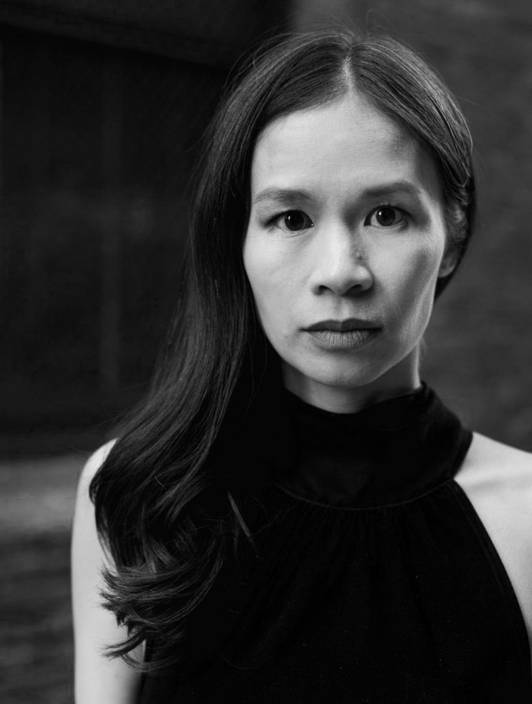 A black-and-white headshot of Aimee, an Asian woman in a dark dress with a "smize" facial expression and medium-long wavy hair over one shoulder