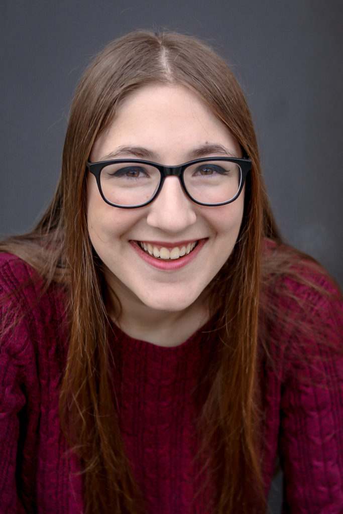 A headshot of Annie, a smiling white person with long straight brown hair, brown eyes, black glasses, and a maroon sweater