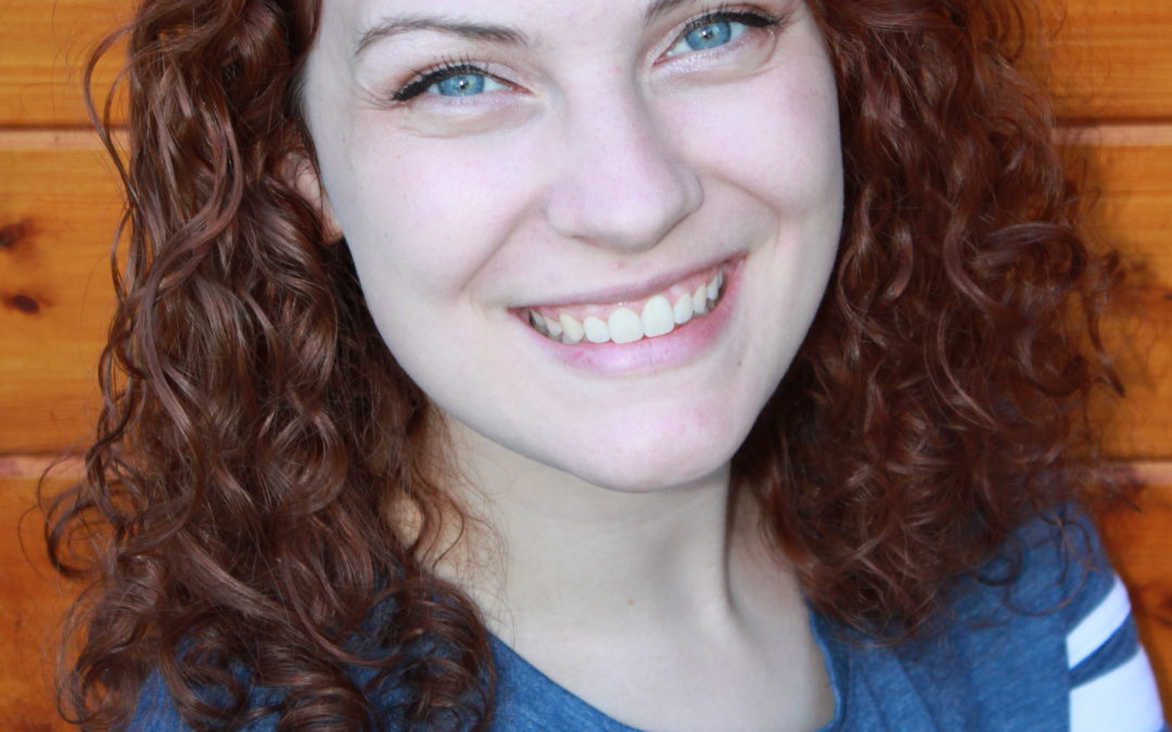 A photo of Karli, a smiling white person with short auburn-colored hair, blue eyes and a blue t-shirt against a wooden background