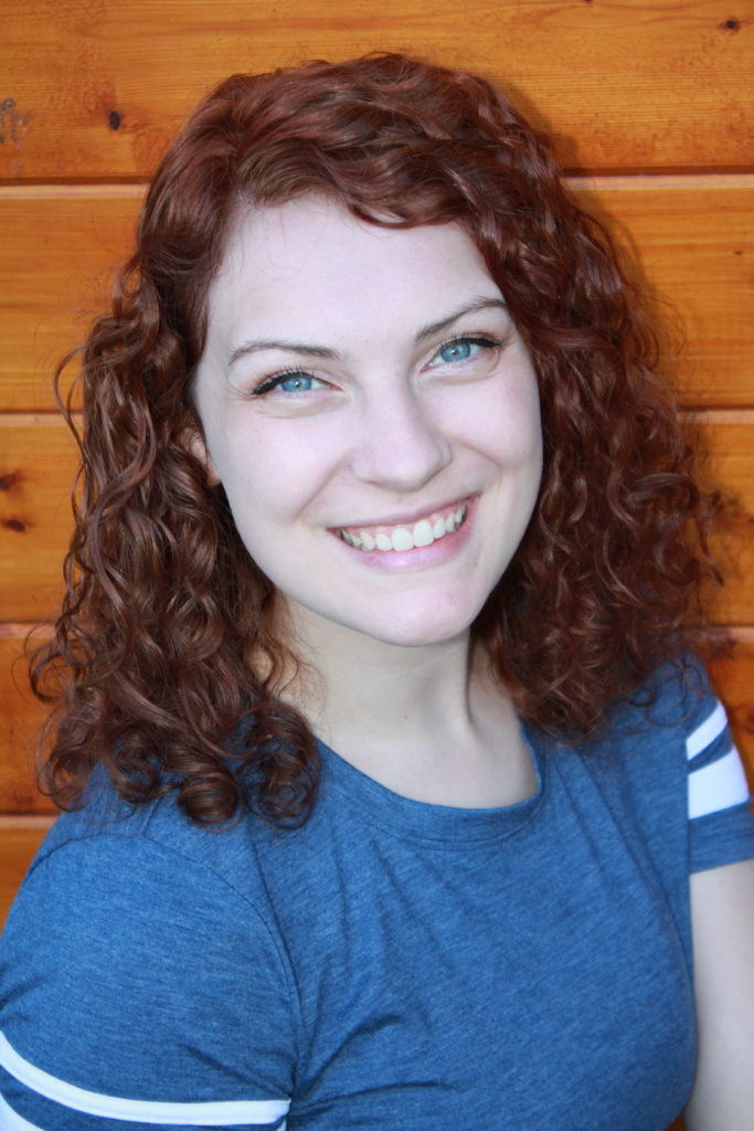 A photo of Karli, a smiling white person with short auburn-colored hair, blue eyes and a blue t-shirt against a wooden background