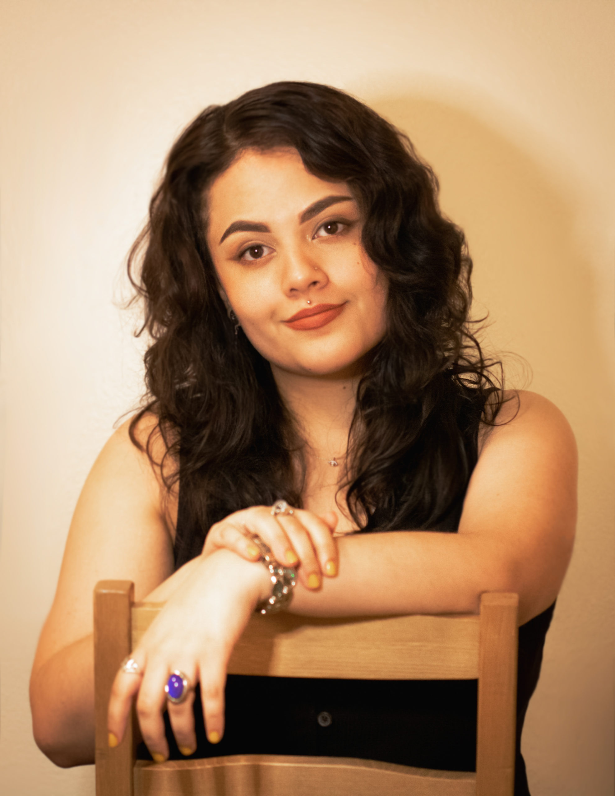 [Image description: a portrait image of Nikeesha, a light-skinned brown woman with brown eyes and curly dark brown hair that falls beneath her shoulders. She is wearing a sleeveless button-up dress and is sitting backwards on a chair with her arms resting on the top rail. End description.]