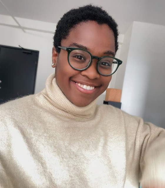 [Image description: headshot image of Zharia, a Black woman with cropped hair wearing glasses with dark green frames, a cream-colored turtleneck, and a smile. End description.]