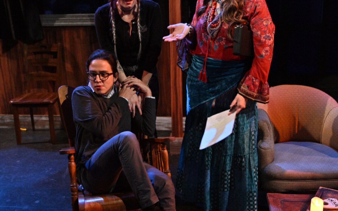 ID: From left: a seated (MERLIN (Phelan Conheady), in a dark sweater and pants, with glasses and ponytail, clutches the hands of BRITTANY (Brittany Rupik), who is standing behind him wearing a black top, with an equally terrified expression. To their left is MEDIUM ( MEDIUM (Jessica Kiely) in a red top and blue long flowing checker-patterned skirt and light-blue turban and holding a piece of paper as she delivers shocking news. End ID.