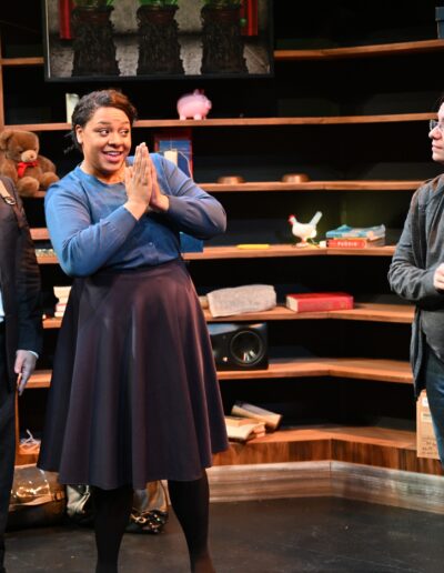 ID: From left: MISSIONARY 2 (Van Lang Pham), wearing a black suit, white blouse, and black tie, standing by MISSIONARY 1 (Talasi Haynes), who is wearing a light violet cardigan and long dark skirt with black leggings, and holding her hands in a prayer position while CALVIN (Kai Winchester) looks onward in a grey top, grey hoodie, and blue jeans. Background: a ramshackle bookshelf. End ID.