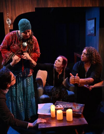 ID: ID: From left: MERLIN (Phelan Conheady), in a dark sweater and pants, with glasses and ponytail, clutches a coffee table as their left hand is gripped by MEDIUM (Jessica Kiely), who is the only one standing. She’s in a red top and blue long flowing skirt and light-blue turban, who also grips the hand of BRITTANY, who is wearing black long-sleeved top and spider web patterned pants and holding the hand of CALVIN (Kai Winchester) who has a “WTF” expression and is in a black top and jeans. Between those two, INTERPRETER (Talasi Haynes) signs the word “with” and wears a black zipper tunic. Background: an antique dark grey TTY teletypewriter lurks in the nook. Foreground: Coffee table with three candles and Ouija board. End ID.