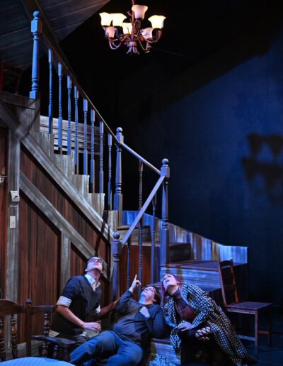 ID: ID: At the base of the staircase, the three roommates, from left, are all staring skyward at something mysterious. Above them is a chandelier and a hint of the attic gables. From left: MERLIN (Phelan Conheady), CALVIN (Kai Winchester), BRITTANY (Brittany Rupik). Merlin is wearing glasses and a dark gray sweater. Calvin is pointing upwards, wearing a grey shirt and jeans. Brittany is wearing a stylishly checkered black-and-white jacket. The scene is lit in a luminous, purple-ish moonlit hue. End ID.