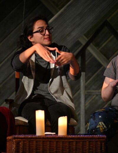 ID: From left: BRITTANY (Brittany Rupik), CALVIN (Kai Winchester), and MERLIN (Phelan Conheady) are in the attic, seated behind a wicker basket and two candles Brittany is wearing a black sweater and dark red corduroy pants. Merlin wears glasses and dark hair tied in a ponytail, plus grey shirt and grey hoodie. Calvin also has glasses, light gray shirt and Baby Yoda pajama pants. All of them are signing “Ouija” in unison. Merlin and Brittany are smiling but CALVIN looks confused. Background: gable pattern with diagonal slats. End ID.