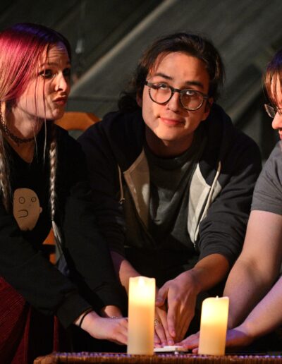 ID: From left: BRITTANY (Brittany Rupik), MERLIN (Phelan Conheady), CALVIN (Kai Winchester), are in the attic staring different directions, fingers on a Ouija planchette waiting impatiently. Brittany is wearing a black sweater and dark red corduroy pants. Merlin wears glasses and dark hair tied in a ponytail, plus grey shirt and grey hoodie. Calvin also has glasses, light gray shirt and Baby Yoda pajama pants. Background: gable pattern with diagonal slats. End ID.