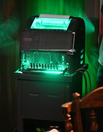 ID: A close-up of a Green Monster TTY teletypewriter, lit up in bright fluorescent green as electric wires stick out and a spool of paper sits on top. It looks like a sinister robot monster. End ID.