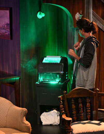 ID: From left: CALVIN (Kai Winchester), MERLIN (Phelan Conheady), and BRITTANY (Brittany Rupik), all standing under a living room alcove. They’re all surrounding a mysterious Green Monster TTY teletypewriter, lit up in a green fluorescent glow that accentuates the keyboard keys. Brittany is wearing a black sweater and dark red corduroy pants. Merlin wears glasses and dark hair tied in a ponytail, plus grey shirt and grey hoodie. Calvin also has glasses, light gray shirt and Baby Yoda pajama pants. Background: painting, New England living room, flashing signal light on wall. End ID.