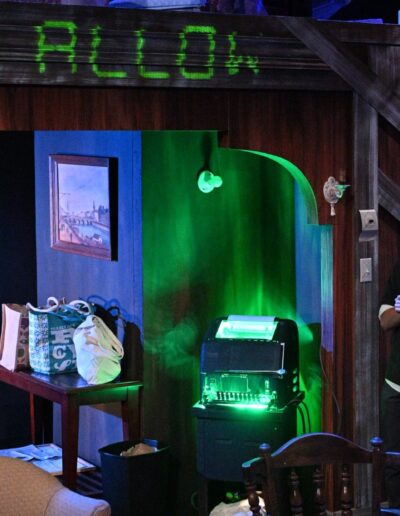ID: Far left: CALVIN (Kai Winchester) stares at the Green Monster TTY with an alarmed quizzical expression. Center: A lit-up Green Monster TTY in neon green lights casting a green backlight to the alcove nook corner. Far right: MERLIN (Phelan Conheady) also staring at the machine in surprise. Above them on the beam, are cut-off captions ending in “... E TO ALLOW.” End ID.