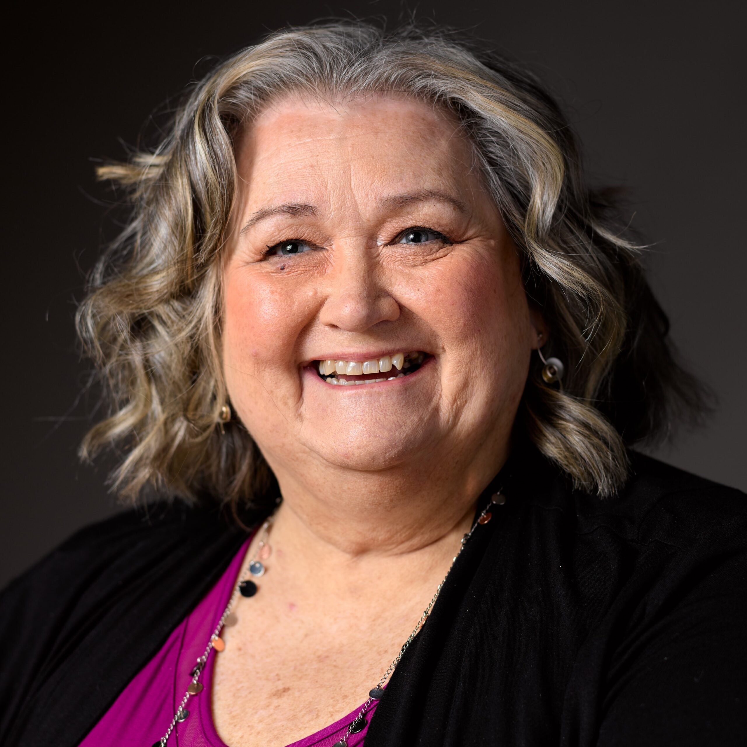 [ID: headshot of Corynn, a round-faced, pale woman with dark brown eyes and chest-length dark brown hair smiles directly into the camera. She is wearing a black cowl-neck shirt. End ID.]