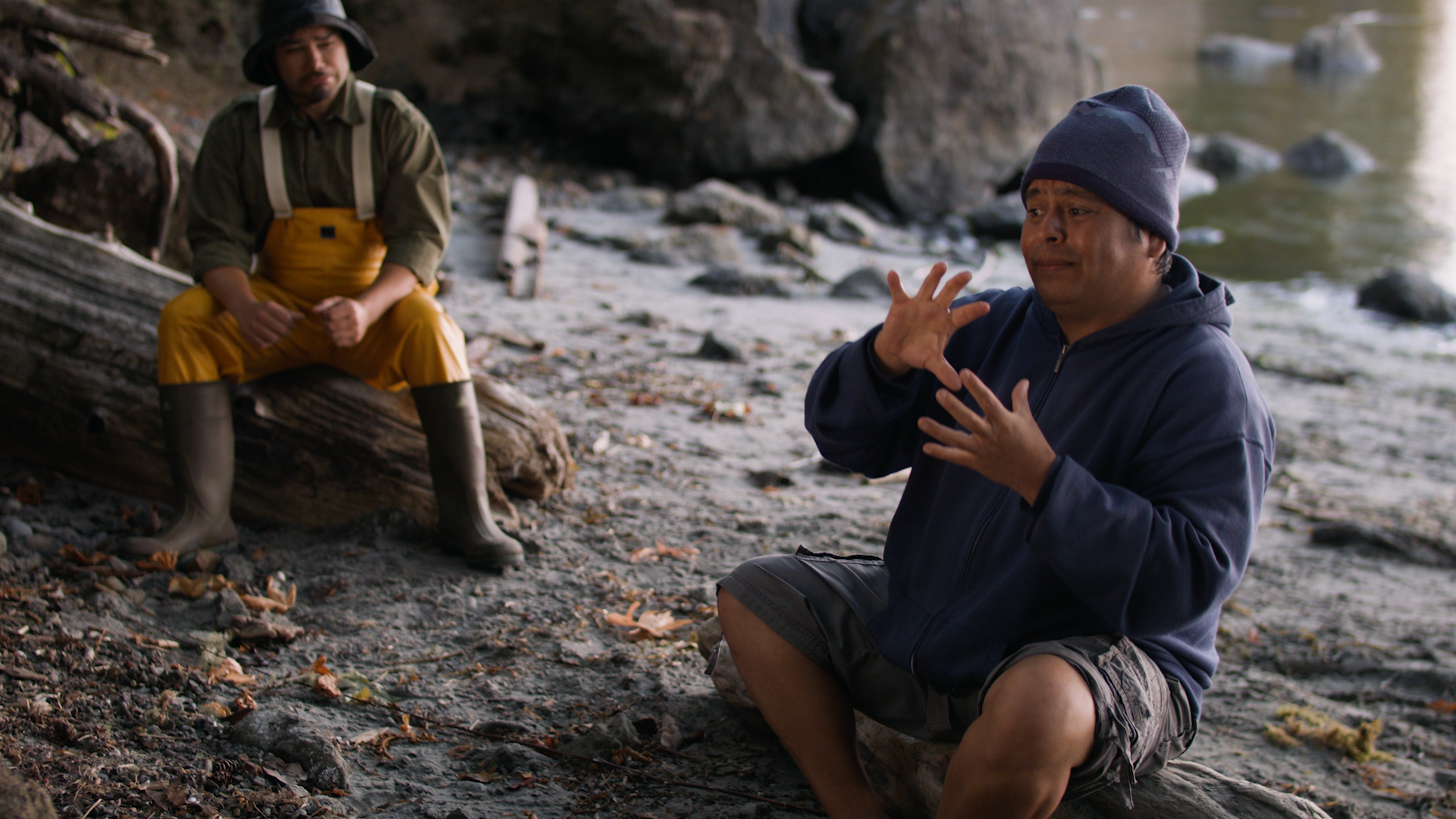 ID: film still from CHANGER: A HAND TELLING. Sonny (played by G. Christian Vasquez) and Johnny (played by Roberto Sandoval) sitting on logs on the beach. They are an indigenous grandfather and grandchild. Sonny is dressed in dungarees and rubber boots, while Johnny is wearing casual clothes. Johnny is signing “Changer” in the foreground as Sonny watches. End ID.