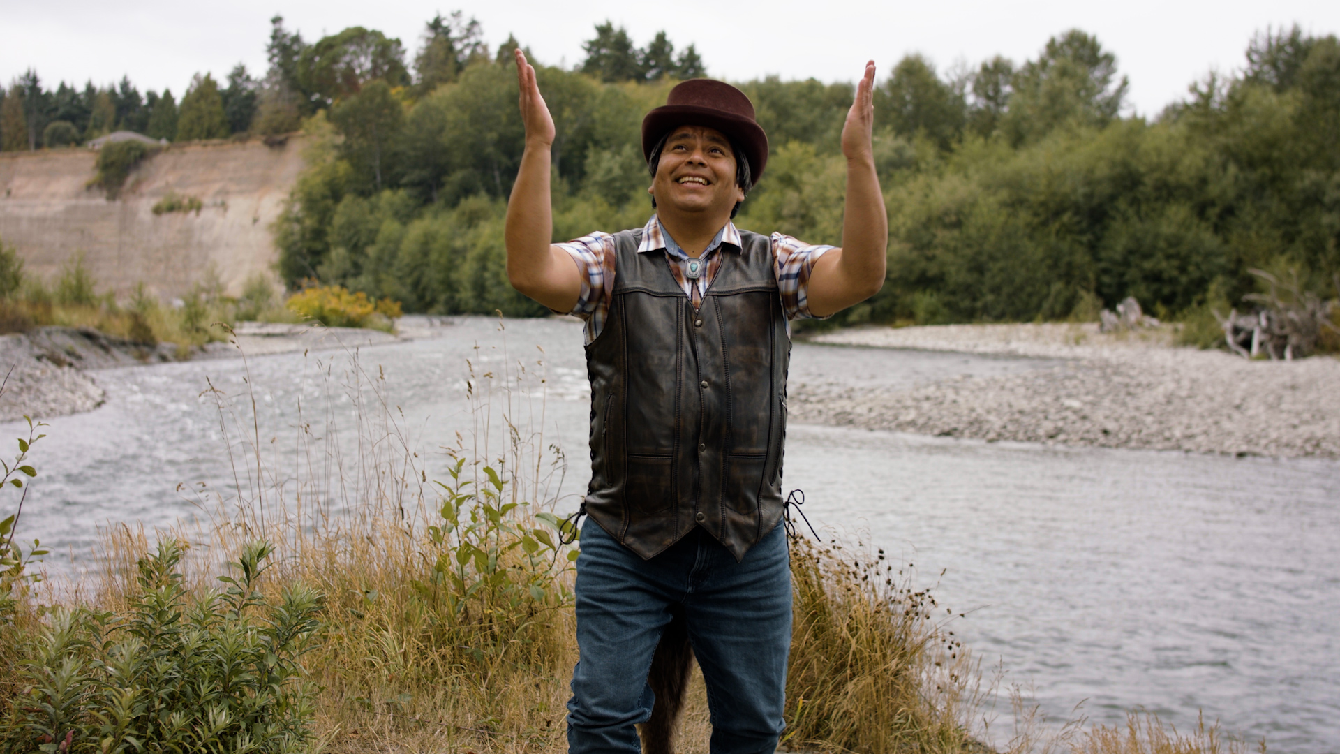 ID: Film still from CHANGER: A HAND TELLING. Coyote (played by Roberto Sandoval), an indigenous person, standing in the grass and sand on the beach. His head is tilted up, and his hands are lifted to the sky. End ID.