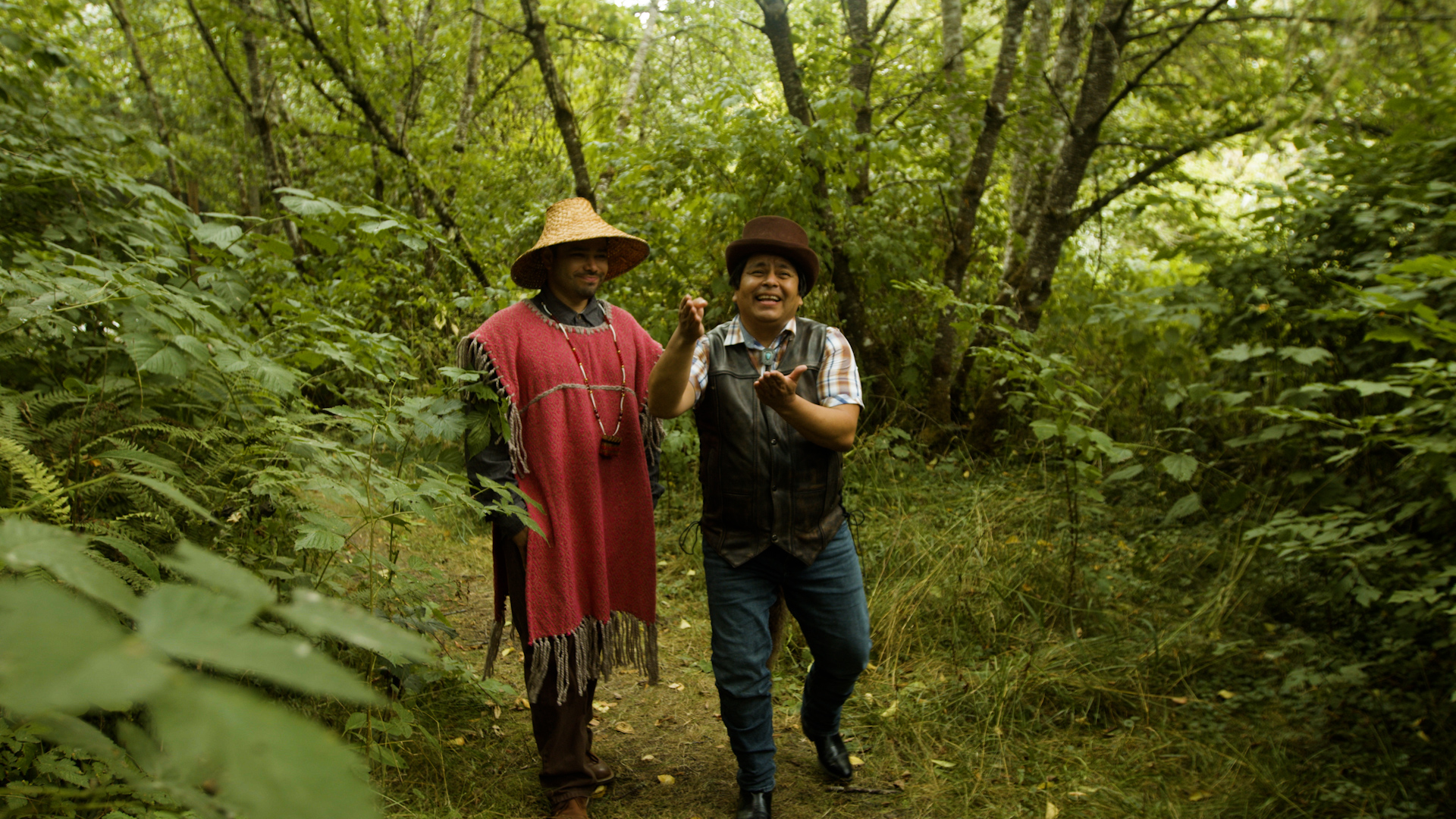 ID: Film still from CHANGER: A HAND TELLING. Changer (played by G. Christian Vasquez) and Coyote (played by Roberto Sandoval), two indigenous people, walking on a forest path full of greenery. Changer is wearing a cedar hat, a red tunic, and a long beaded necklace. Coyote is wearing a brown top hat, a brown vest, a plaid shirt, and a bolo tie. Coyote is signing “huckleberry pie” in ASL, as Changer watches. End ID