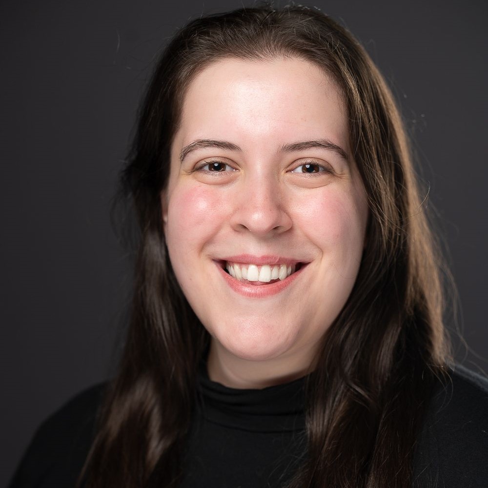 [ID: headshot of Corynn, a round-faced, pale woman with dark brown eyes and chest-length dark brown hair smiles directly into the camera. She is wearing a black cowl-neck shirt. End ID.]