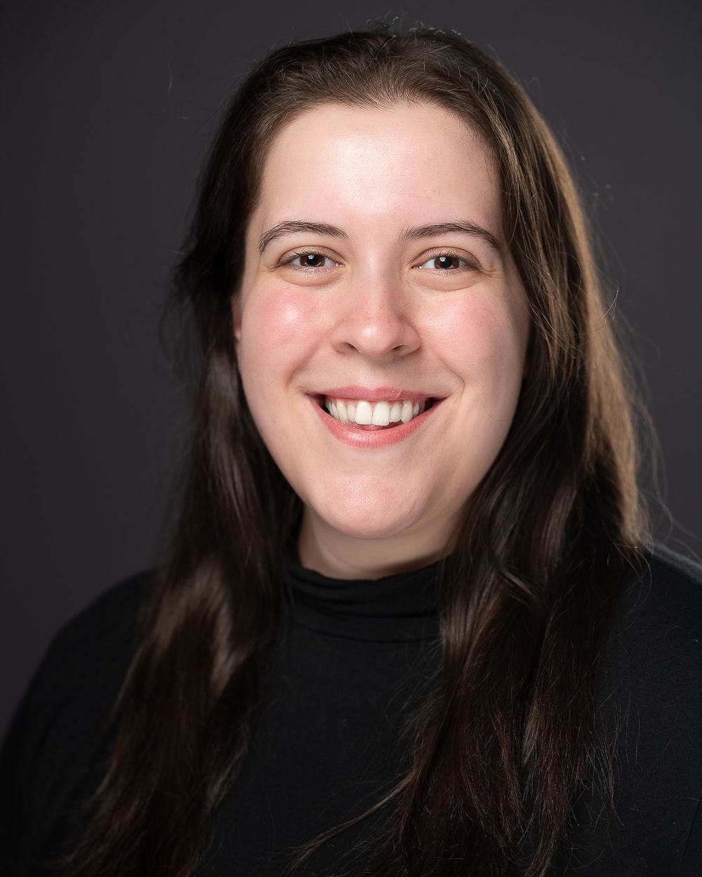 [ID: headshot of Corynn, a white woman with long, straight brown hair worn loose and brown eyes. She's smiling brightly and wearing a black cowl-neck shirt. End ID.]