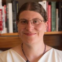 [ID: headshot of Corynn, a white woman with long, straight brown hair worn loose and brown eyes. She's smiling brightly and wearing a black cowl-neck shirt. End ID.]