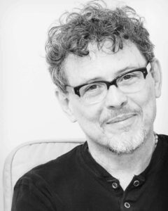 [ID: black-and-white headshot of playwright José Rivera, pictured here smiling gently with cropped curly hair, a trimmed beard, dark-colored henley shirt, and semi-rimless glasses. End ID.]